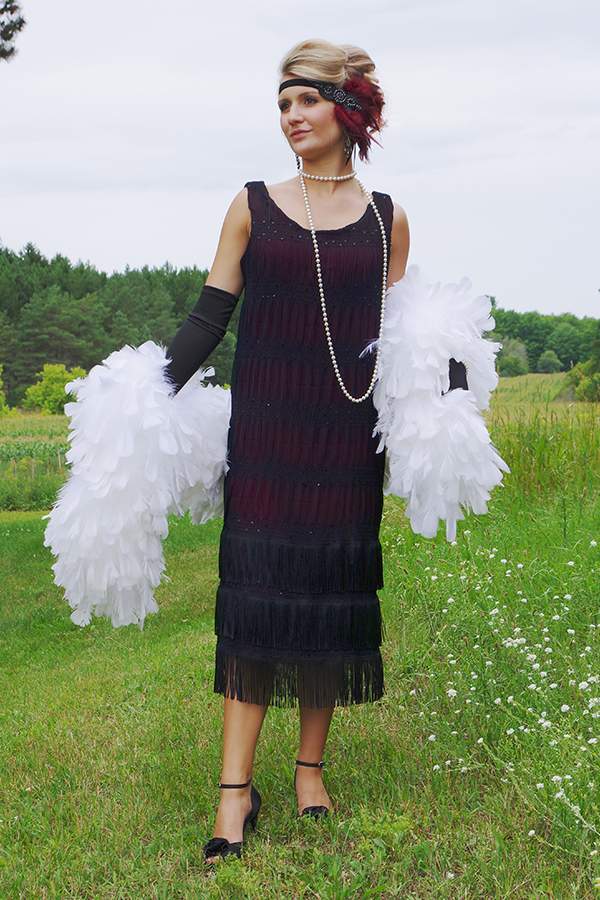 Blonde woman wearing black flapper style dress with a large white boa standing in a field. 